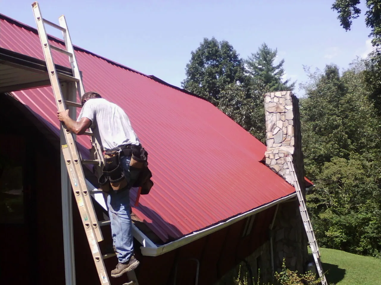 Roofer on a ladder beside a red metal roof and stone chimney — Soffit & Fascia Repair by Roofing Brothers in Staatsburg